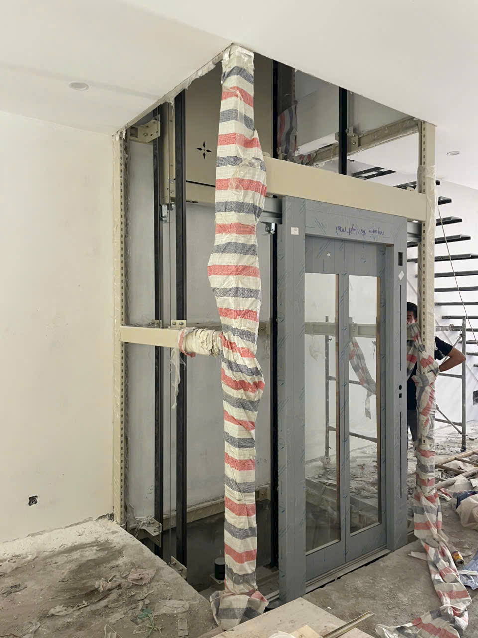 Thang máy gia đình đang lắp đặt trong nhà, khung và cửa kính xám còn bọc bảo vệ